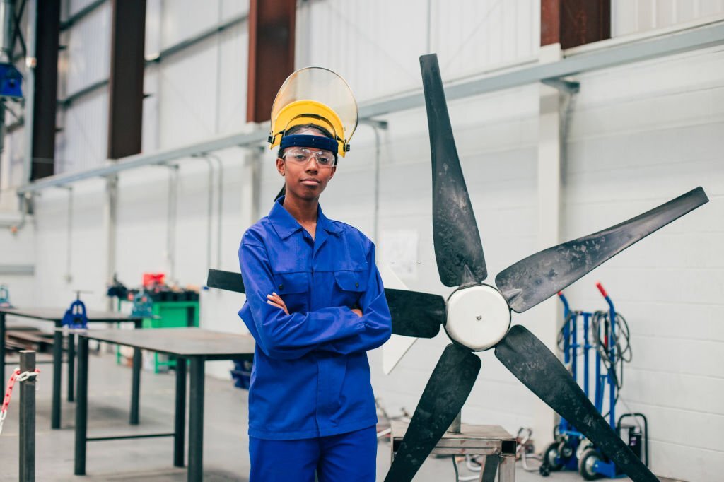 A young woman stands and looks at the camera in an engineering workshop. She is wearing a protective mask and eyewear as well as blue coveralls.